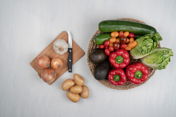 Vegetable basket with tomatoes, red peppers, zucchini, avocados, and salad. Wooden cutting board with fresh garlic, onion and potato, kitchen knife on white background