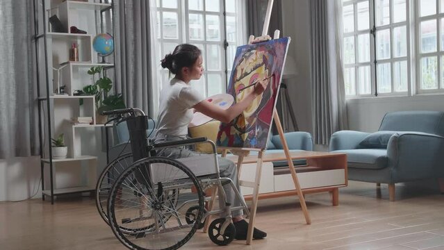 Side View Of An Asian Artist Girl In Wheelchair Holding Paintbrush Mixed Colour And Painting A Girl On The Canvas
