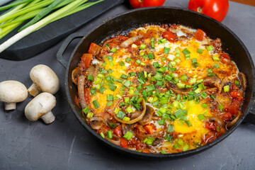 Shakshuka in a frying pan on a gray background next to mushrooms, tomatoes and green onions.