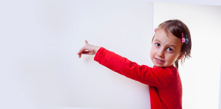Cute little child girl behind white empty banner. She pointing to white wall. Free copy space area for text or design.