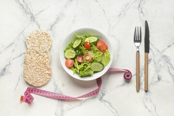 Bowl of healthy salad, crispbreads, cutlery and measuring tape on light background