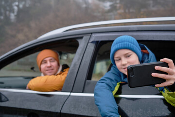 Happy father and son Sitting together in a new car on a journey. Family are resting on the side of the road on a road trip. Child takes pictures on smartphone. Happy family travels.
