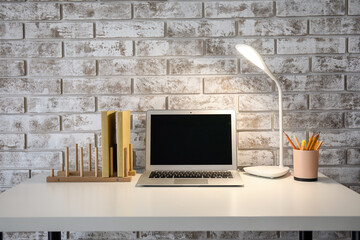 Wooden organizer with books, glowing lamp and laptop on table near brick wall