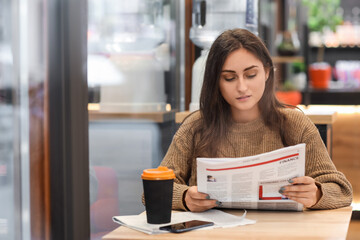 Obraz premium Pretty young woman reading newspaper at table in cafe