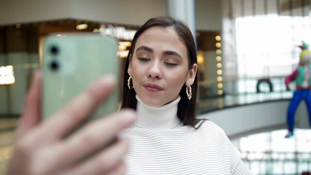 A girl in a white jacket and long hair makes a selfie on a smartphone. A photo of yourself in a mall. A beautiful young girl admires herself on her phone screen.