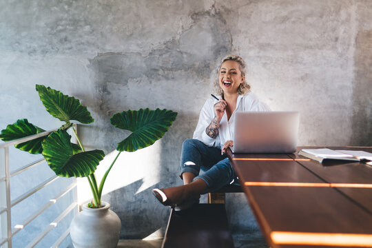 Portrait of happy blonde hipster girl with modern laptop computer smiling at camera during remote working, joyful female freelance copywriter with digital netbook technology laughing at desktop