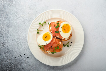 Sandwich with delicious toppings on top: smoked salmon, eggs, herbs and microgreens radish, black sesame seeds over white plate on concrete background. Healthy open sandwich superfood. Top view.