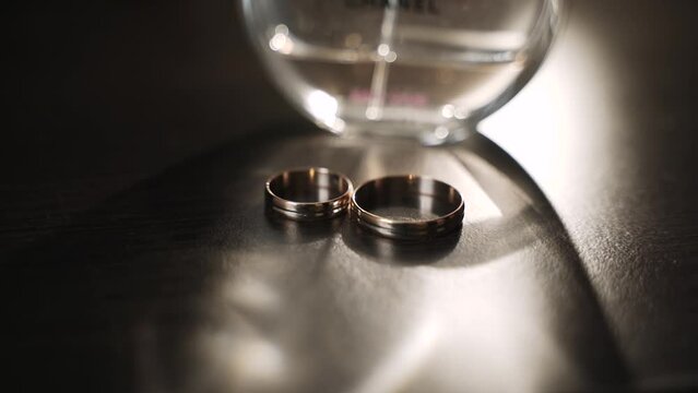 Bimetal wedding rings on a wenge-colored tabletop under a shifting beam of light with a shadow from a perfume bottle. 