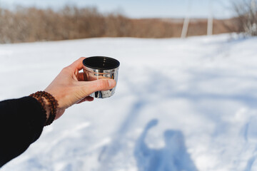 A hand holds a cup of tea against the backdrop of a winter forest. Hot tea in a mug, an iron glass, the concept of drinking hot drinks in nature. Hiking in the mountains