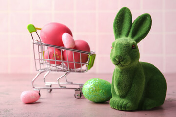 Cute Easter bunny and shopping trolley with painted eggs on pink background