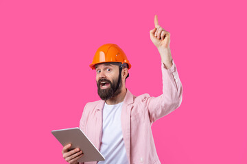Portrait of young construction engineer wear orange hard hat, in a pink jacket standing on red studio background. A man with a tablet in his hands.