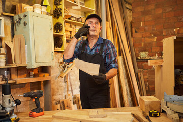 Portrait of a senior carpenter looking at blueprints plans  to make a piece of furniture and talking on the phone with a client in the carpentry workshop