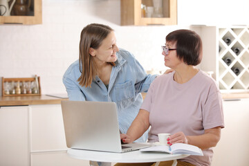 young woman teaching senior mother to use internet on laptop at home. daughter helps her elderly mother figure it out online with her personal account, teaches at modern gadget indoors.