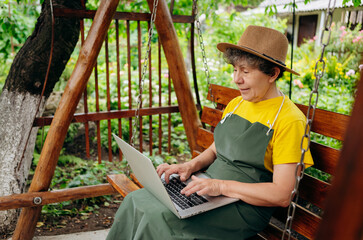 Obraz na płótnie Canvas Senior woman gardener in a hat is watching something on a laptop computer and making a video call in the yard while outdoors.