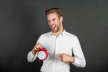 Happy crazy guy sticking tongue while pointing finger at alarm clock dark background, timing