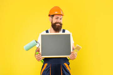 happy man in helmet and work clothes hold paint roller brush and blackboard with copy space