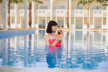 Young likable pretty woman in pink swimwear, drinking yummy fresh cocktail, while swimming in outdoor luxury blue resort pool. Tropical vacations concept