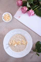 Breakfast in the cafe. A plate of oatmeal porridge on a light background. Greeting card and pink rose on a light background. An empty space for the inscription on the postcard. Vertical photo.