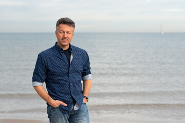 Outdoor portrait handsome mature man walking at the beach. Attractive adult male model posing at seaside in blue jeans and shirt.