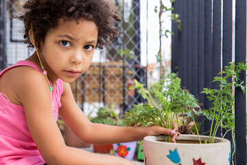 Lovely mixed race latin girl sitting on the balcony and looking at camera while planting an organic vegetable in a pot in a sunny day.  Springtime and nature concept.