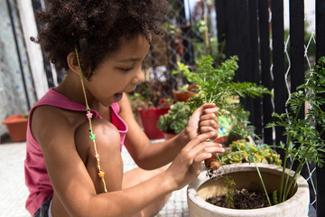 Happy mixed race curly hair girl sitting on the balcony focused on taking an organic carrot from a pot in a sunny day. Springtime and nature concept.