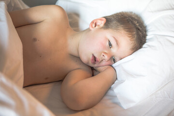 Kid 7 year old lying on bed, Sleepy child waking up the morning in his bed room with morning light, sad boy lies on a bed in the room
