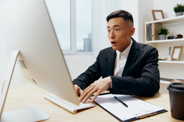 Fototapeta premium Asian man in a suit glass of coffee sits at a tablein front of a computer Lifestyle