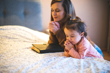 Mother and her daughter reading from bible and praying in their knees near the bed - 491915194