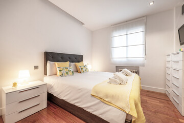 Bedroom with a king size bed with a yellow bedspread, matching cushions and folded towels, a chest of drawers with lots of drawers in a vacation rental apartment