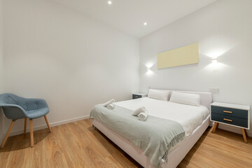 Fototapeta na wymiar Bedroom with a king size bed with a white duvet, white bedside tables with blue drawers and a blue chair with wooden floors in a short-term rental apartment