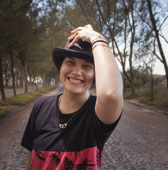 Fototapeta premium Young male gender fluid female on a cobblestone street with a tunnel of trees in the background wearing a black top hat. Mention the lgbt theme, gender change, sexual freedom and sexuality in youth.