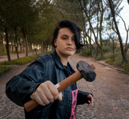 Young male gender fluid female on a cobblestone street with a tunnel of trees in the background holding a sledgehammer. Mention the lgbt theme, gender change, sexual freedom and sexuality in youth.