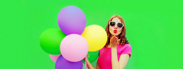 Portrait of young woman with bunch of colorful balloons blowing her red lips sending sweet air kiss on green background