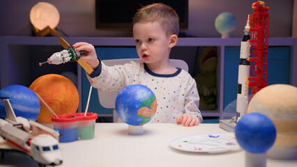 Little blonde boy kid plays with Sputnik from constructor, flies near planets of solar system in evening. Child imagines himself as pilot of spaceship. Desire fly in space, flying on spaceship.