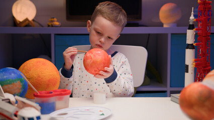 Blond boy paint planet solar system Mercury with colorful paint sitting home table in evening,...