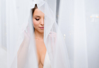 Close view of thoughtful bride, which wearing in sophisticated wedding dress with plunging decolletage, covering head and naked body by veil, looking down while posing in room during bridal morning