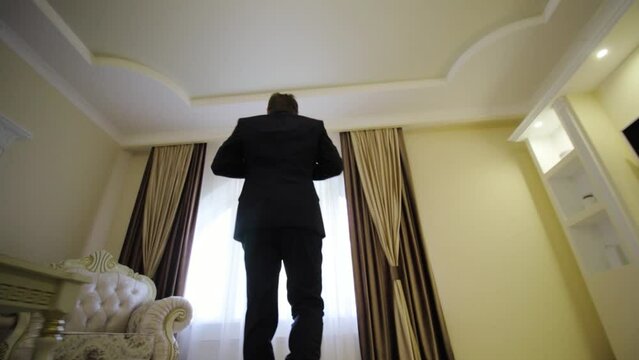 Man in black suit walks across hotel room to curtained window. Groom fastens the button on his ja
