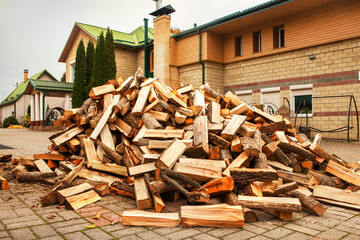 A pile of split firewood for heating the house, unloaded in the yard, against the backdrop of the house, natural heating sources.