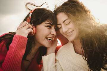 Tender couple of young girlfriends carefree outdoors listening and sharing music from headphones -...