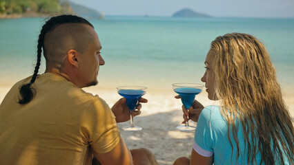 The love couple is holding a glass of blue curacao cocktail, on sea. Man with plait and blonde...