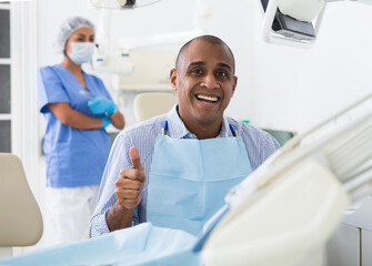 Portrait of satisfied hispanic man in dental office showing approval gesture with thumbs up after treatment