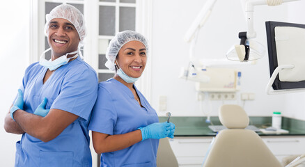 Two professional hispanic dentists in blue uniforms standing back to back in modern office of dental clinic