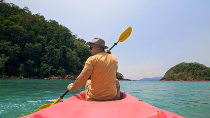 Young man with sunglasses and hat rows pink plastic canoe along sea against green hilly islands...