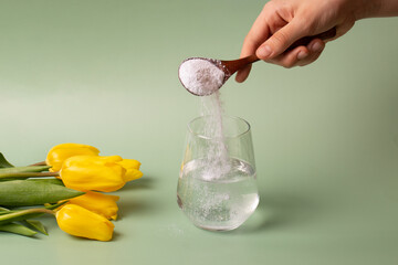 The girl with her hand pours white collagen powder with a wooden spoon into a transparent glass of...