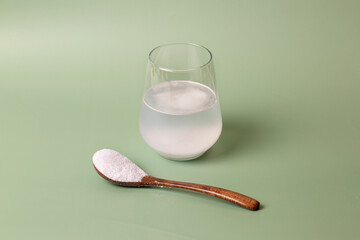 White collagen powder in a transparent glass with water and a wooden spoon on a green background.