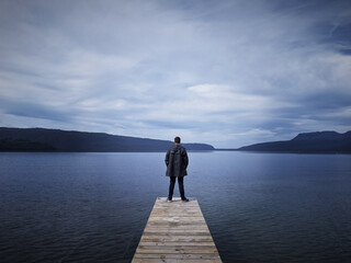 Lone man standing at the end of a wooden pier overlooking a lake under overcast skies