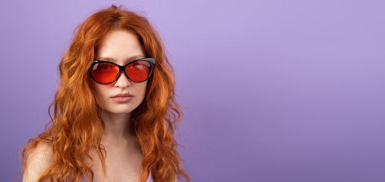 Pretty young woman with red hair wearing sunglasses for the summer time posing on purple background with copy space.