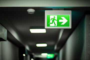 Green fire exit sign on hallway aside of smoke alarm device. Fire prevention and building safety background. - obrazy, fototapety, plakaty