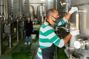 Portrait of male winery worker in mask controlling winemaking process, checking wine near metal tanks for wine fermentation