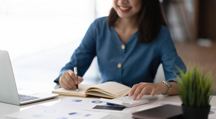 Cheerful young woman with laptop and jar and writing in notebook while sitting at table and working on freelance project at home
